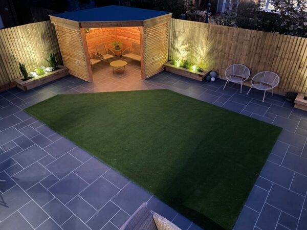 A compact garden with dark paving, a central lawn and an illuminated timber garden room.
